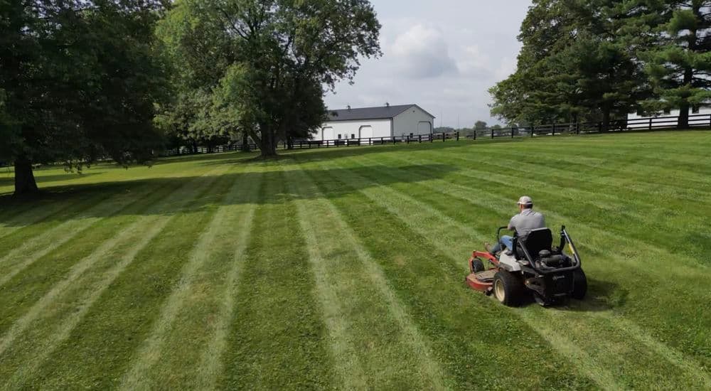 Man mowing grassy field with striped pattern and farmhouse in the background.