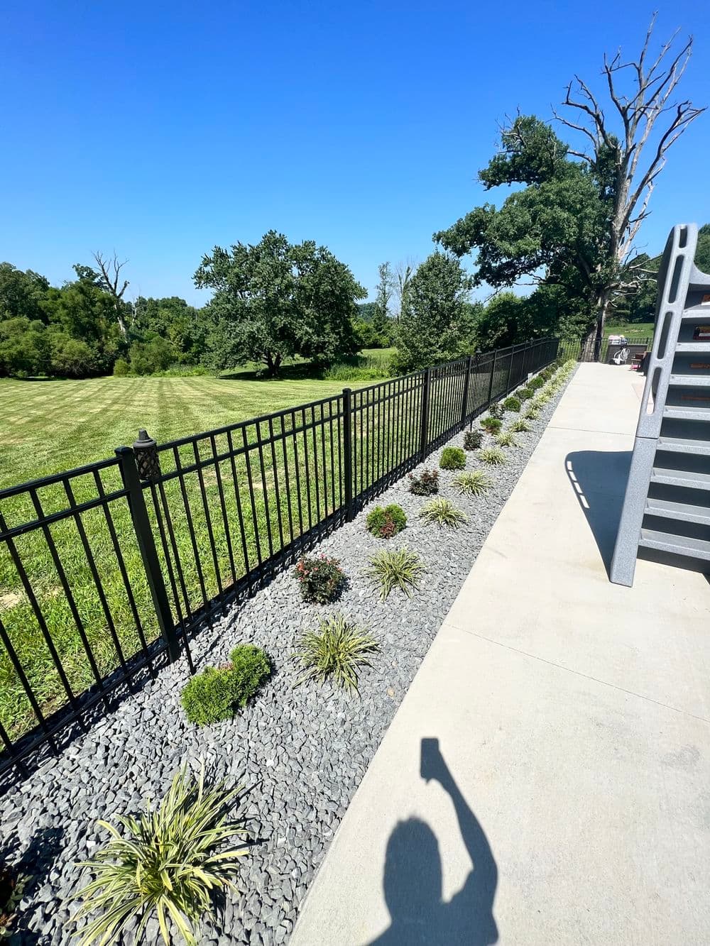 Black metal fence lines a landscaped yard with greenery under a clear blue sky.