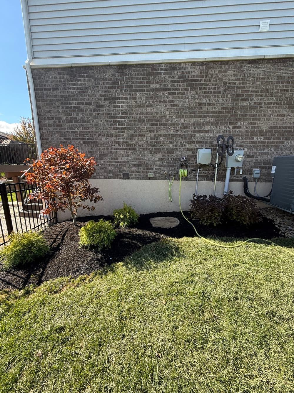 Landscaped yard featuring shrubs, mulch, and a hose beside a brick home.